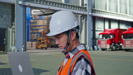 close up side view of asian male engineer with safety helmet working on a laptop while standing , outside of logistics distributions warehouse
