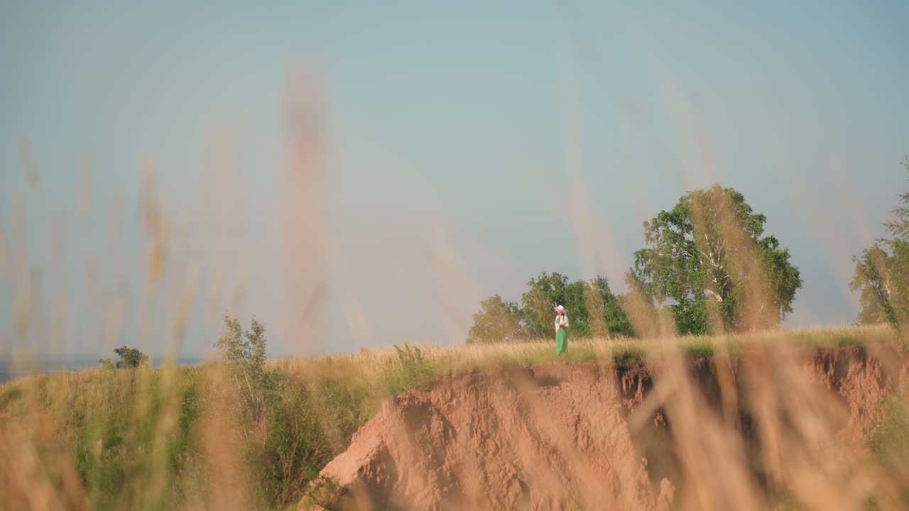 distant woman in green outfit and white hat approaches cliff edge surrounded by tall grass and forest trees, observing steep red terrain below under clear sky in warm sunny summer countryside