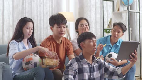 asian teenagers cheering, eating and watching football game on tablet at home