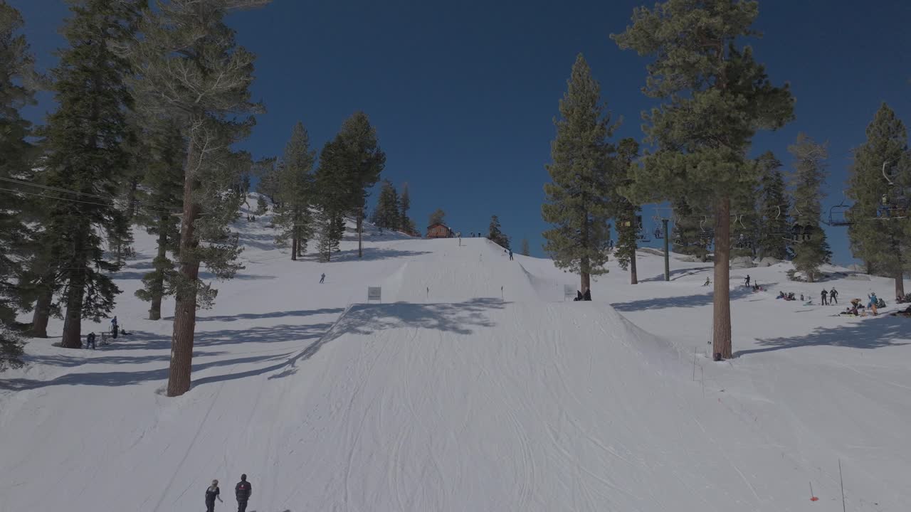 Serbest stil atlayışları ve rampaları olan kayak parkı, havada numaralar yapan kayakçılar ve snowboardcular, izleyen seyirciler, yamaçta görünen kabin, uzun gölgeler oluşturan çam ağaçları, masmavi gökyüzü, güneşli hava
