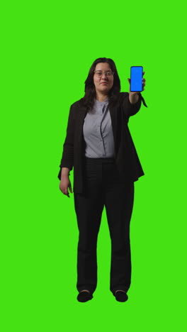 vertical video full length shot of businesswoman holding blue screen mobile phone towards camera standing against green screen 1