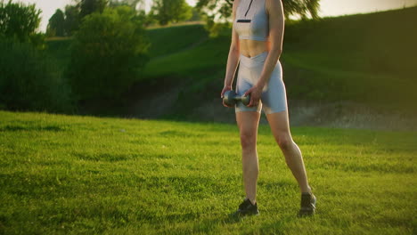 a young woman in sportswear leans forward with dumbbells in nature in a park at sunset. workout. work on a beautiful body in the morning or at sunset