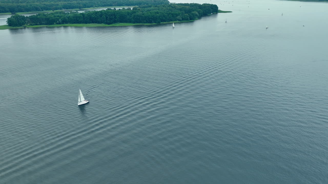 Aerial shot of a sailboat navigating through wide-open calm waters, with a distant forested island in the background under a serene sky