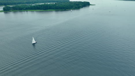 aerial shot of a sailboat navigating through wide-open calm waters, with a distant forested island in the background under a serene sky