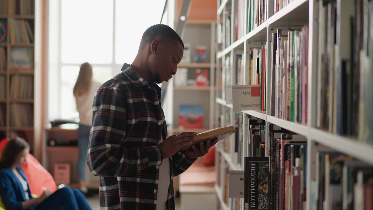 lector masculino pasa por un libro de pie al lado de una estantería. joven investigador afroamericano da la vuelta a las páginas buscando información para un proyecto científico. biblioteca universitaria
