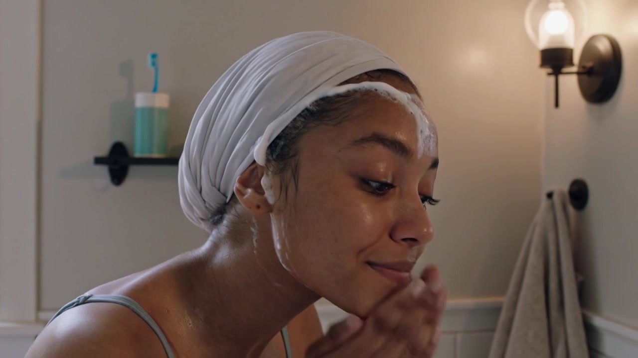 Mulher com turbante e regata aplicando espuma, massageando e enxaguando o rosto, limpando-o na penteadeira, pia.