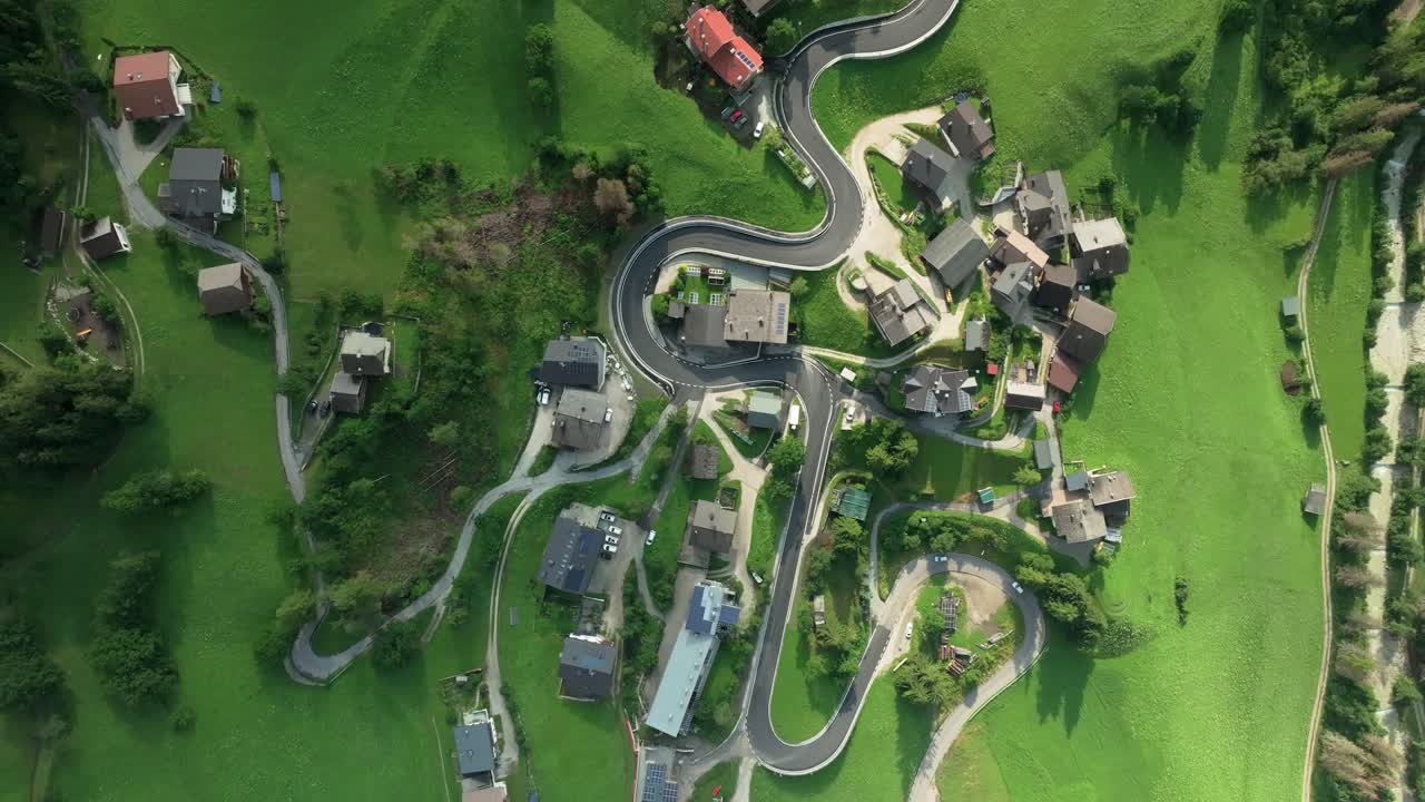 Overhead aerial tilt-down view captures cars navigating a winding road in the quaint village of La Val, South Tyrol, Italy. Lush green hills flank the picturesque scene. Camera moves up. LuPa Creative