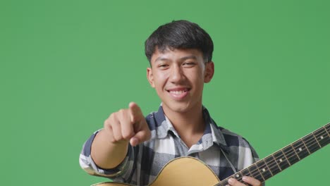 primer plano de un adolescente asiático con una guitarra haciendo un gesto honesto y sonriendo en el fondo de la pantalla verde