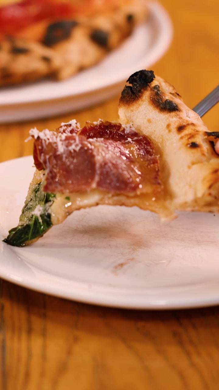 Inquadratura ravvicinata di mani che usano posate per posizionare una fetta di pizza al prosciutto su un piatto.