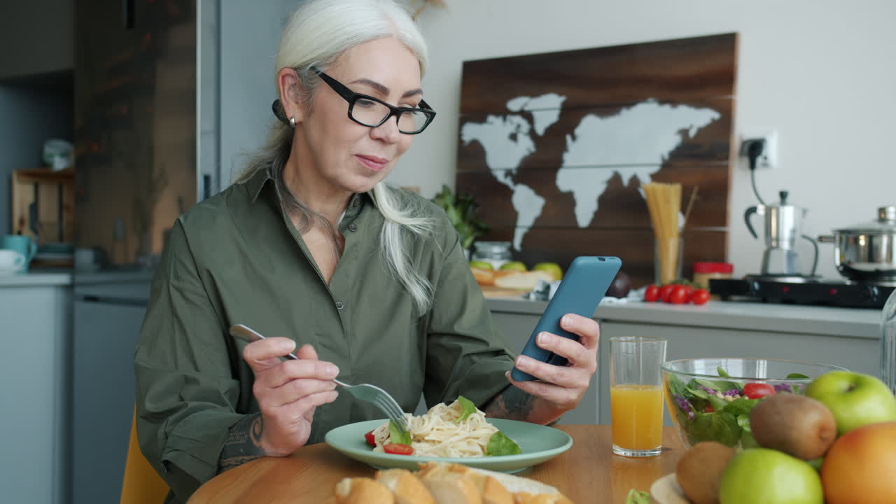 Senior Woman Eating Pasta and Using Smartphone in Kitchen
