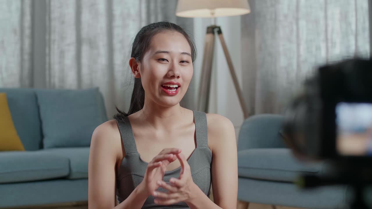 젊은 아시아 트레이너 여성은 스포츠 옷을 입고 집에서 가르치는 운동을 녹음하는 동안 손을 흔들고 카메라에 말합니다.