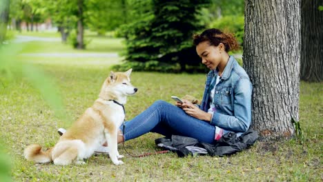 side view of pretty mixed race girl using smartphone relaxing in park under tree while her cute shiba inu dog is sitting near her owner and enjoying nature.