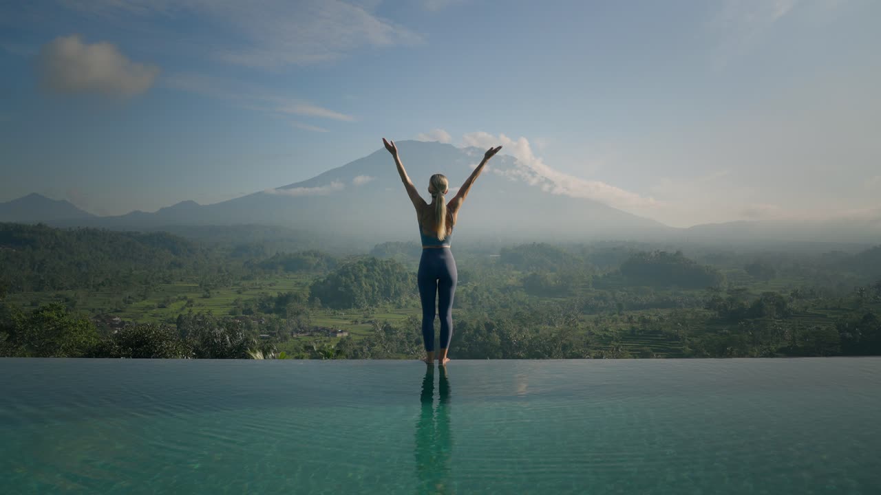 atractiva mujer europea rubia en el borde de la piscina infinita que se extiende en una pose de yoga de saludo ascendente, monte agung, amanecer