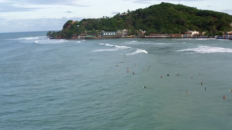 Rotating-aerial-drone-shot-of-large-group-of-surfers-resting-in-the-water-waiting-for-the-next-wave-on-the-famous-tropical-destination-Pipa-beach-in-Rio-Grande-do-Norte,-Brazil-on-a-summer-day