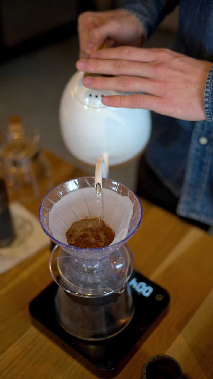 Close up of a man brewing pour-over drip coffee in a slow, circular motion on a wooden table. Vertical