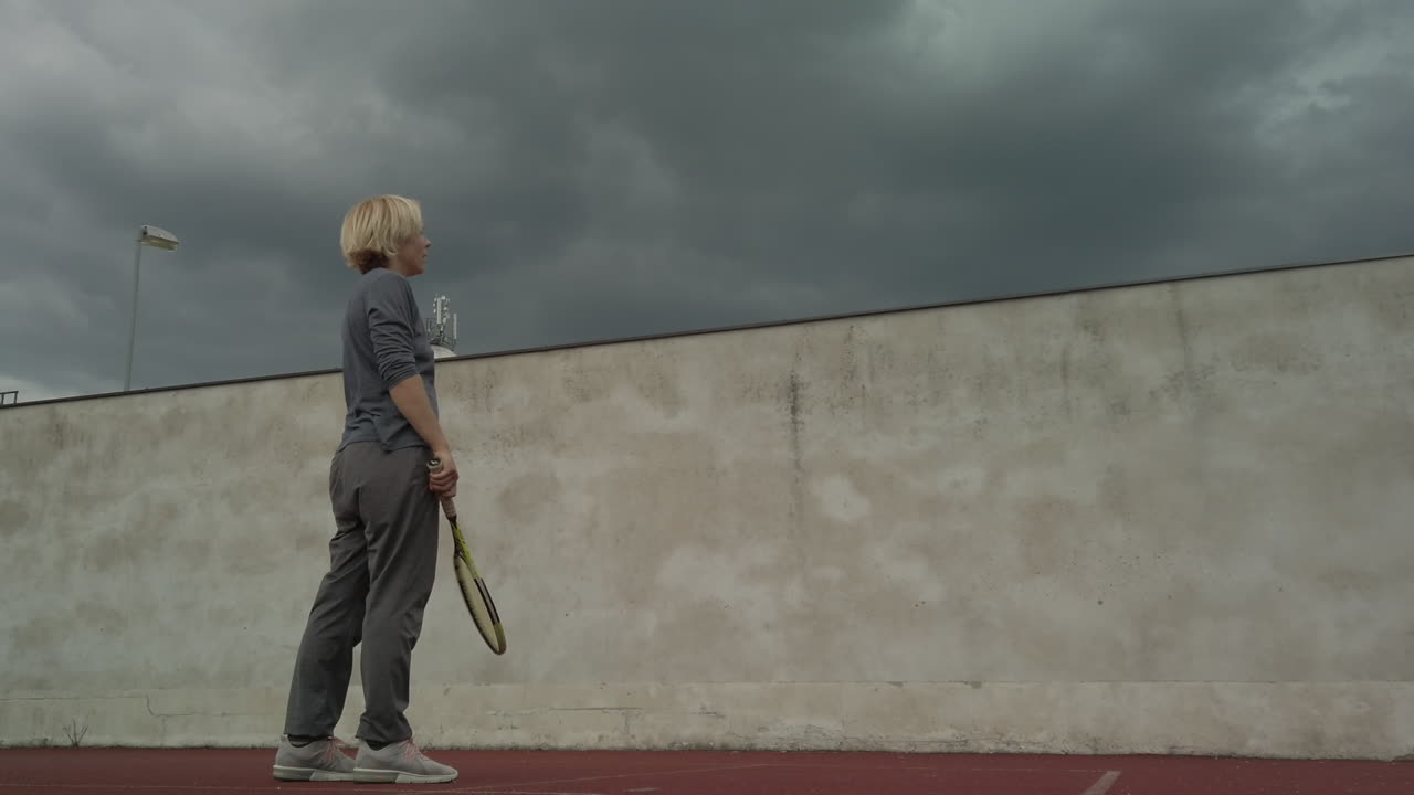 Una donna si allena a tennis contro un muro mentre si addensano nuvole temporalesche, inquadratura dal basso