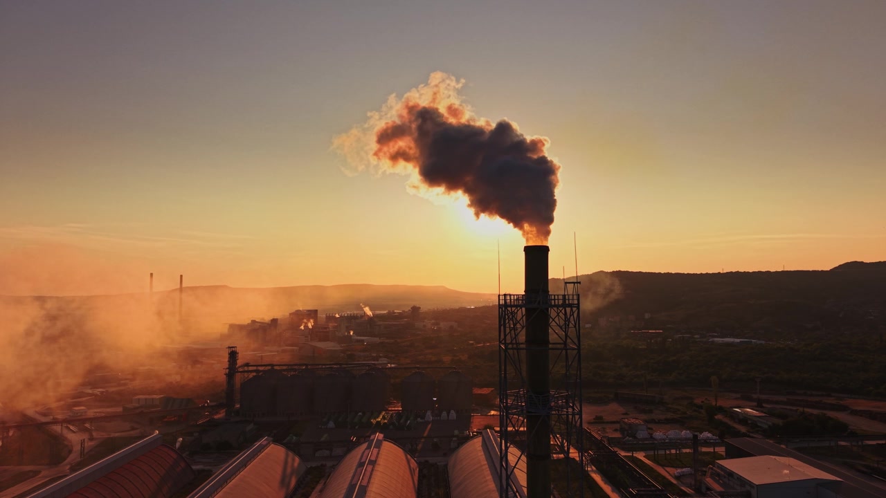 Fumo che sale da una ciminiera di fabbrica al tramonto, vicino a un'area industriale.