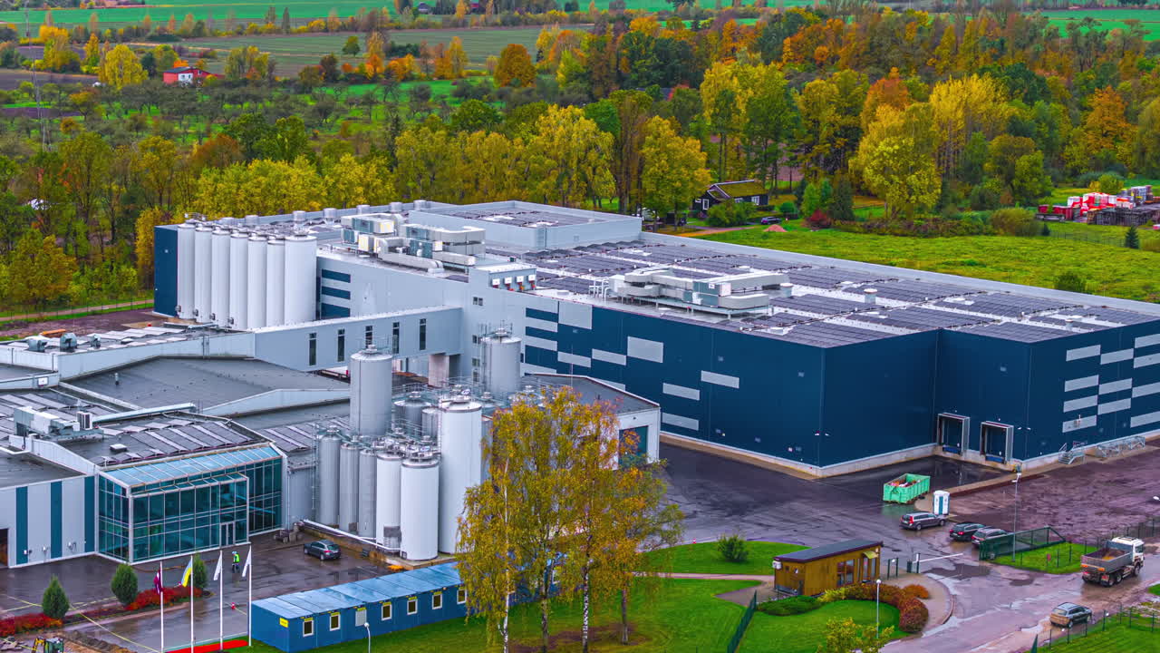 Drone timelapse of a large industrial factory with storage silos and modern blue warehouse, surrounded by autumn forest and farmland. Corporate manufacturing, logistics, and infrastructure