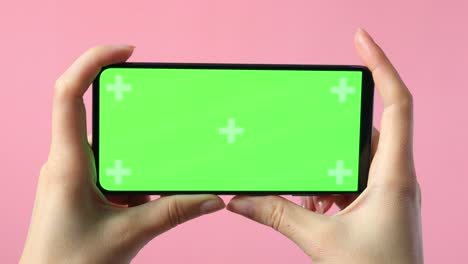 close-up of female hands holding smartphone with blank green screen showing copy space for app or website design, on pink background , mockup.