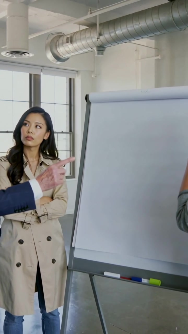 Video verticale: Persona che indica una donna in cappotto beige verso un flip chart durante una riunione in un loft