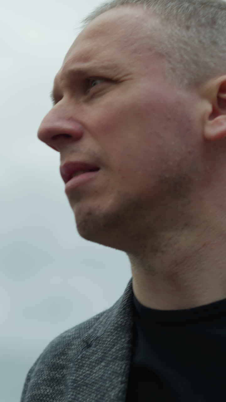 A close-up view of an aged white man wearing a grey suit and a black polo shirt, looking around with a serious expression