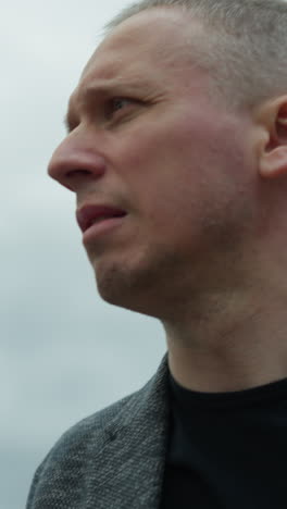 a close-up view of an aged white man wearing a grey suit and a black polo shirt, looking around with a serious expression