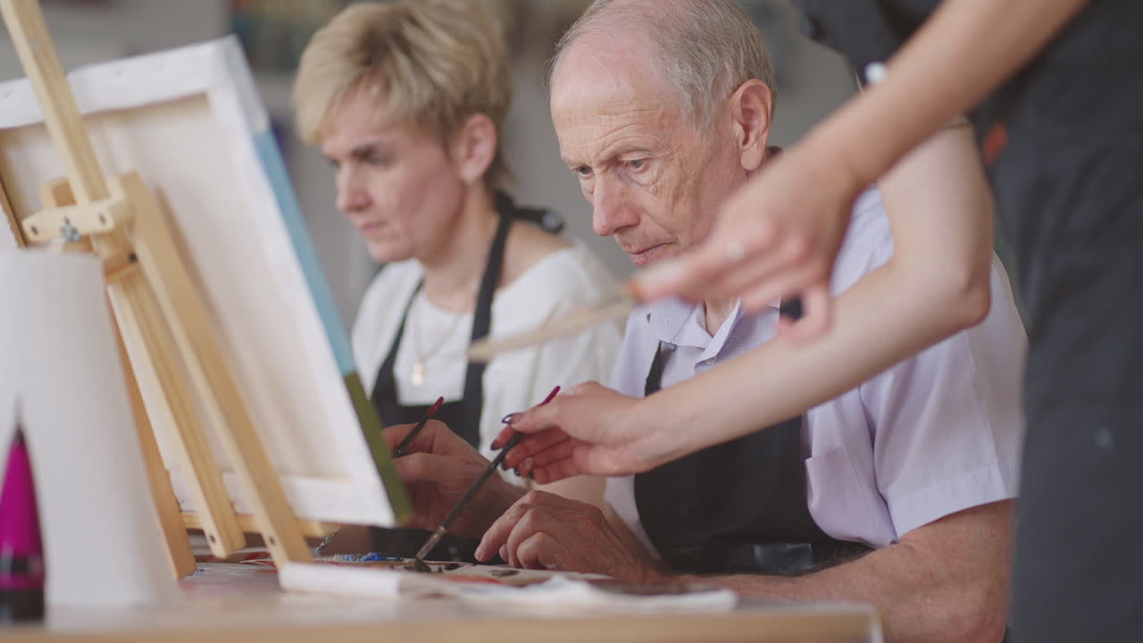 A young woman teacher helps an old pensioner learn to paint a picture. Senior man learning to draw