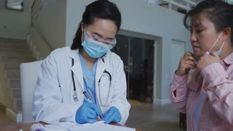 Asian-female-nurse-wearing-face-mask-and-gloves-writing-during-consultation-with-female-patient