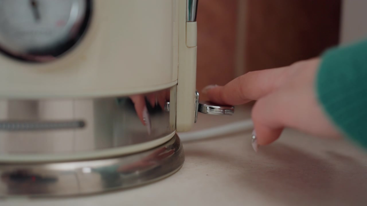 Closeup kettle switch being turned by small hand on new apartment countertop, blue LED indicator glowing, stainless base and cord visible, cozy movein morning routine, intimate domestic scene