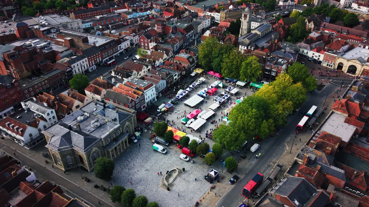 Ripresa aerea con drone sulla Piazza del Mercato di Salisbury durante una giornata di mercato affollata con bancarelle, acquirenti ed edifici storici, con la Cattedrale di Salisbury visibile sullo sfondo. Filmato nel Wiltshire, Inghilterra
