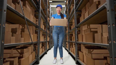 full body of asian female courier in blue uniform showing a carton to camera and smiling while delivering it in warehouse