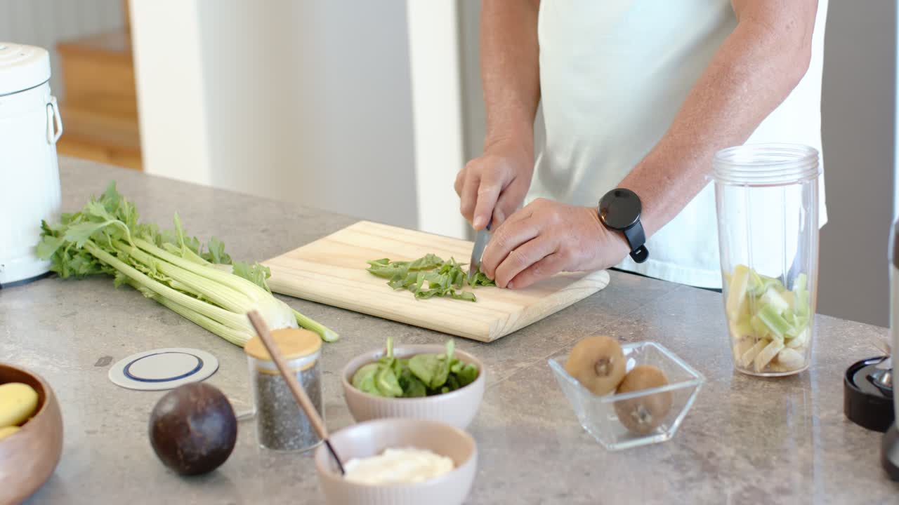 Manojo de verduras de hoja verde siendo colocado mientras se pican ingredientes en una tabla de cortar en la encimera de la cocina para una ensalada.