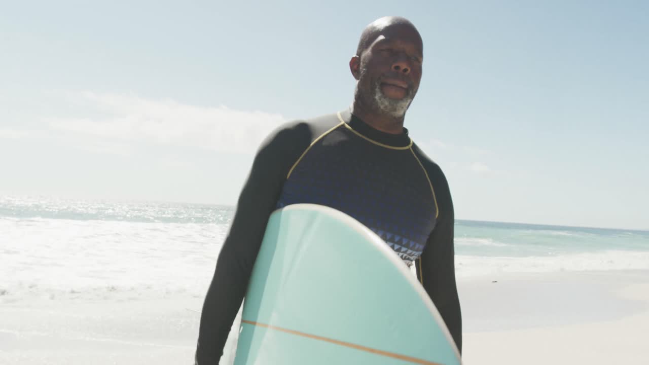 Uomo anziano afroamericano che cammina con tavola da surf su spiaggia soleggiata