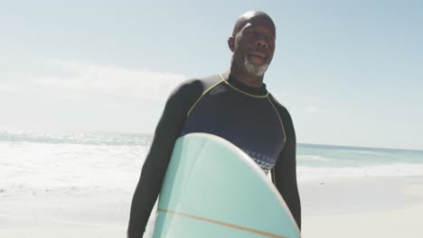 Hombre-Afroamericano-Senior-Caminando-Con-Tabla-De-Surf-En-La-Playa-Soleada