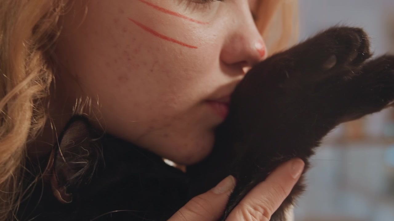 Close up of woman with red face paint holding and petting black cat focusing on their interaction. scene shows intimate bond between them in soft indoor lighting emphasizing texture details