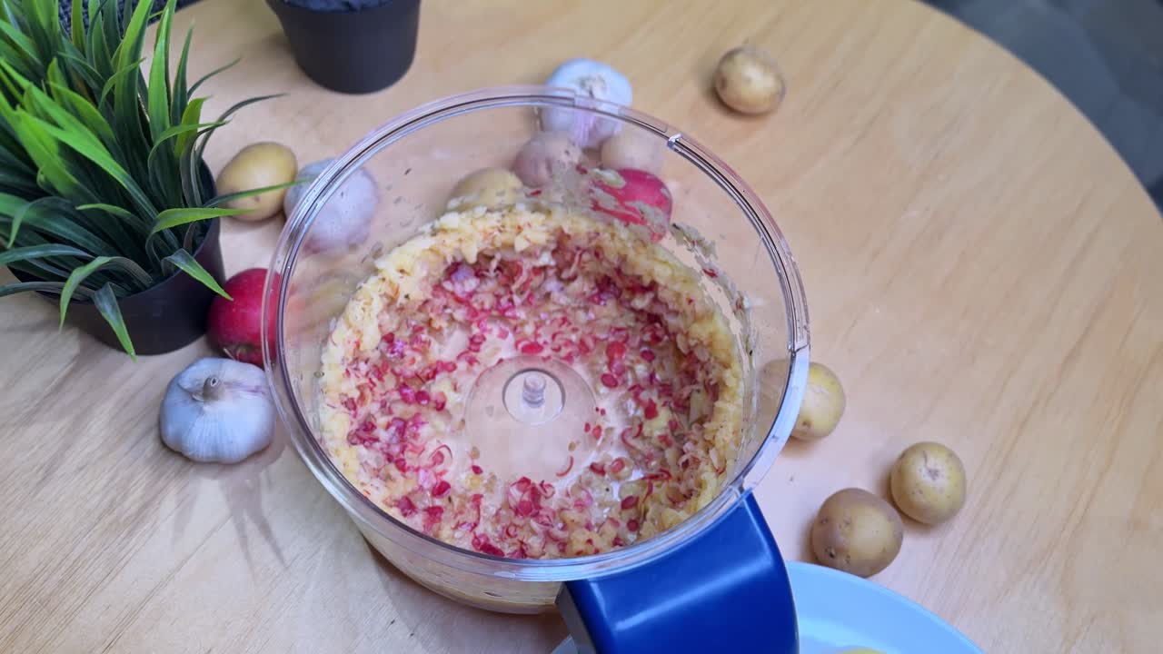 Overhead close-up of a food processor bowl with minced garlic, onion and chili mix on a wooden table, surrounded by potatoes and whole garlic for home cooking prep