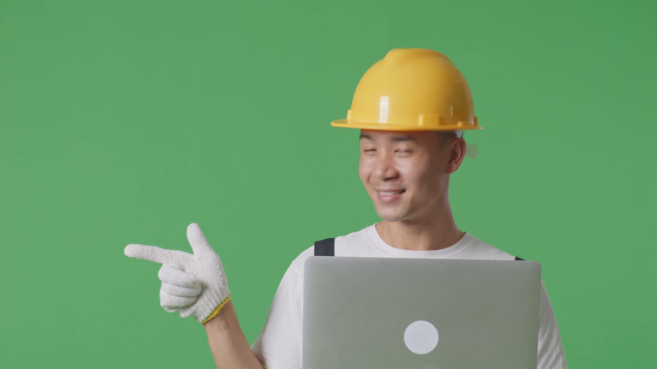 Close Up Of Asian Man Painter Wearing Safety Helmet Using A Laptop And Pointing To Side While Standing In The Green Screen Background Studio