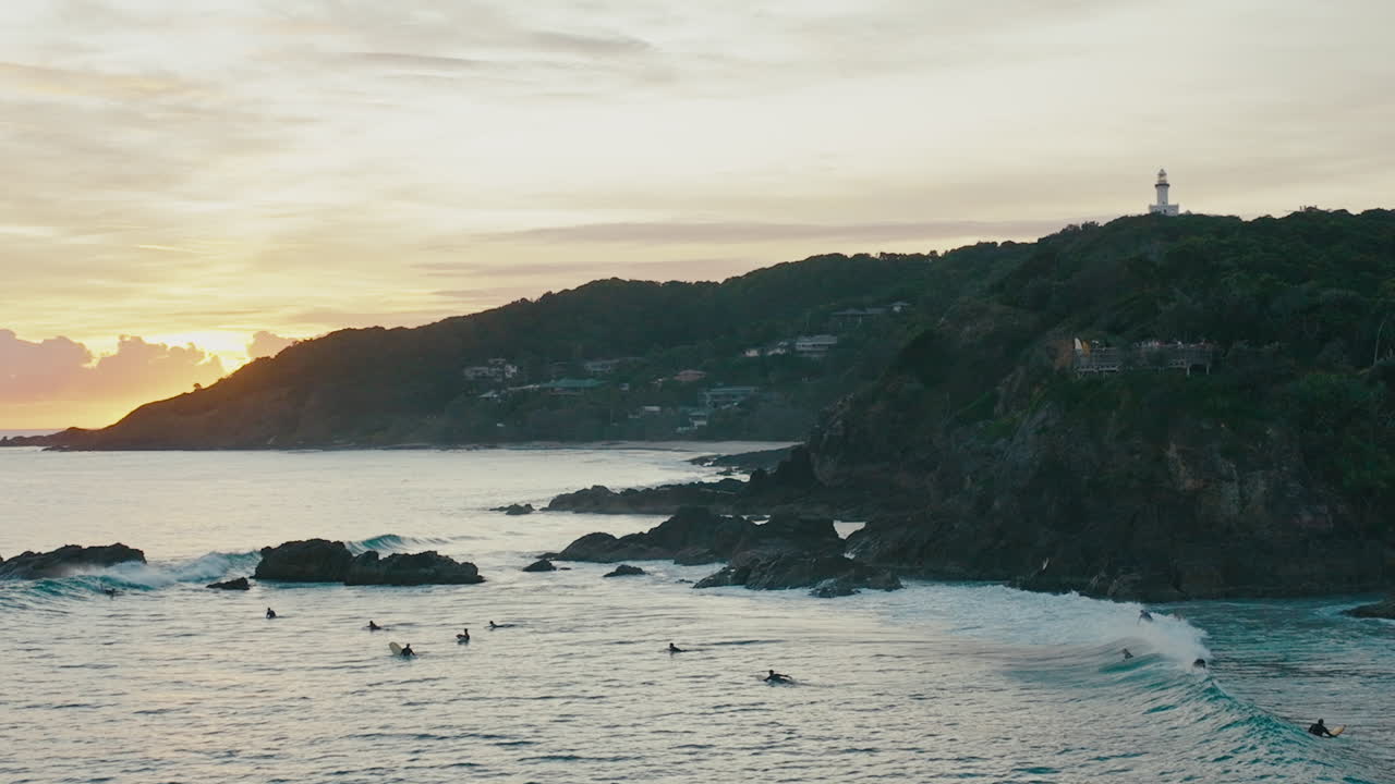 La luce della sera illumina la costa rocciosa di Byron Bay mentre le onde lambiscono dolcemente la costa; faro sullo sfondo e surfisti che cavalcano le onde, vista aerea