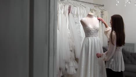 beautiful-girl-looking-wedding-dress-in-the-cabin-compared-to-other-dresses.-Preparing-for-the-wedding.-Buying-a-wedding-dress.-The-seamstress-inspects-the-finished-custom-made-wedding-dress.