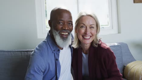 portrait of happy senior diverse couple in living room sitting on sofa, embracing and smiling