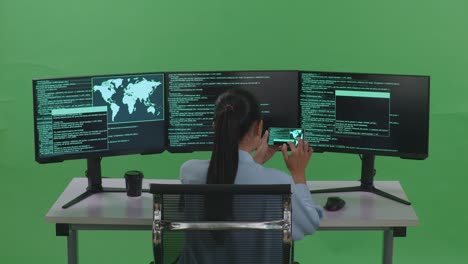 vista trasera de una desarrolladora asiática trabajando con un teléfono móvil mientras escribe código con múltiples pantallas de computadora en un estudio de pantalla verde