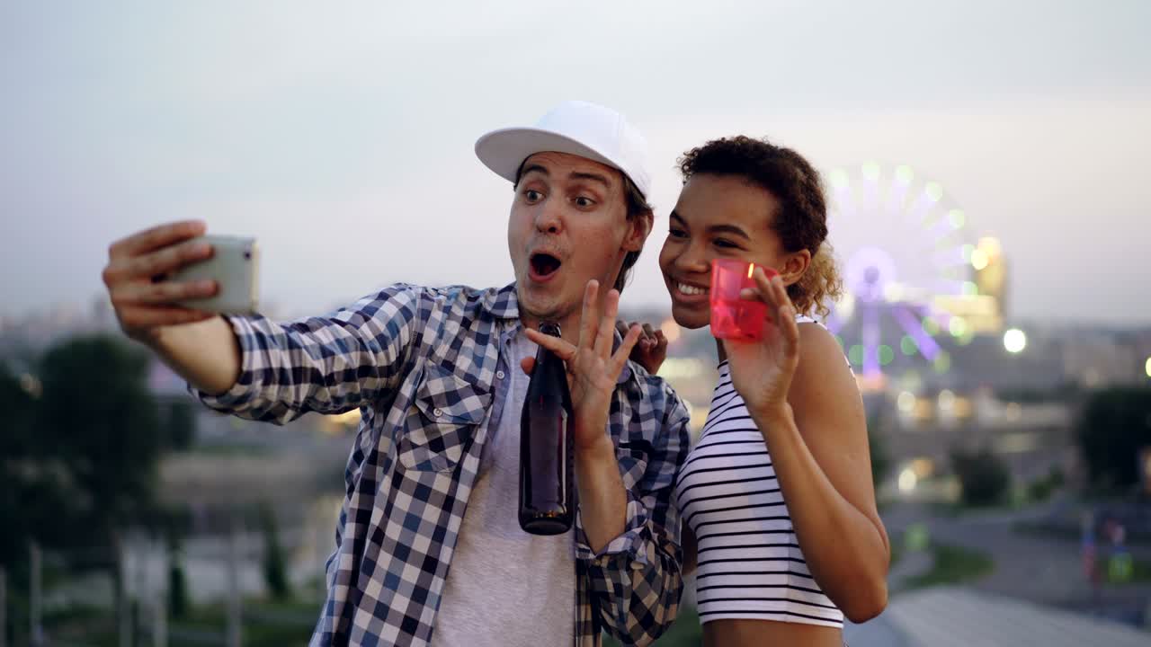 amigos jóvenes atractivos con rostros expresivos se están tomando selfies con teléfonos inteligentes en la azotea sosteniendo bebidas en vasos y botellas.