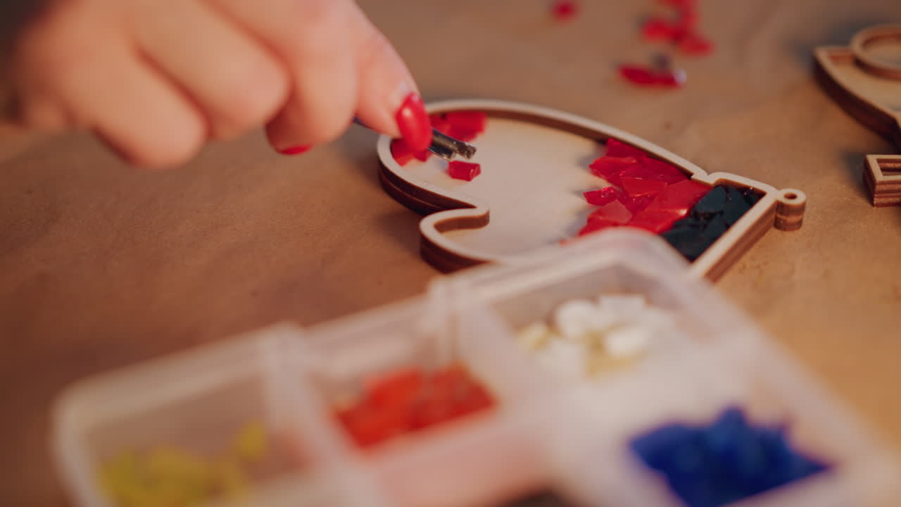 Closeup hands filling heart ornament with red beads, mosaic pattern forming inside wooden frame, tweezers placing tiny pieces with steady motion, warm glow and intimate festive atmosphere, final