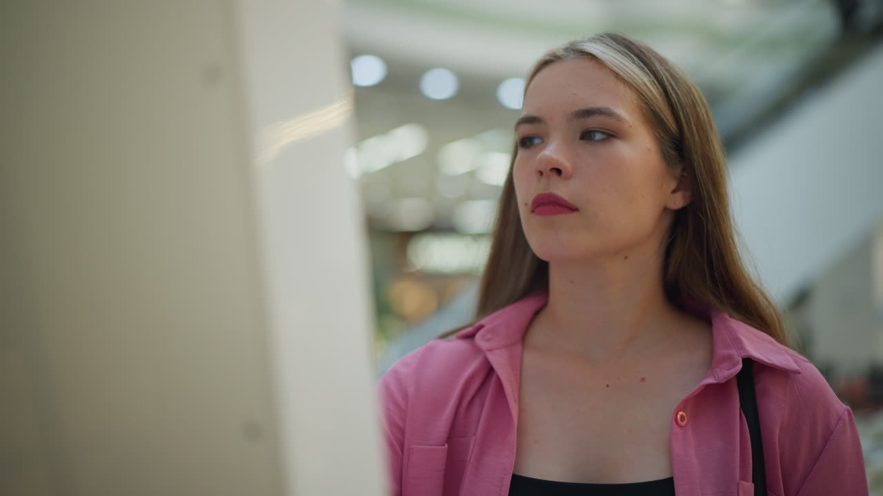 dama de vestido rosa con bolso negro intensamente enfocada en el uso de una pantalla en un centro comercial bien iluminado, el fondo presenta luces borrosas y escaleras mecánicas, y parece profundamente concentrada en su tarea