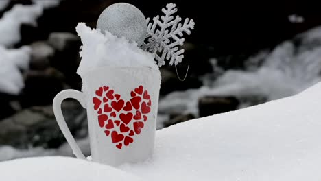 taza blanca con un diseño de corazones rojos lleno de nieve, una esfera navideña plateada y un copo de nieve, con un arroyo en el fondo de la naturaleza