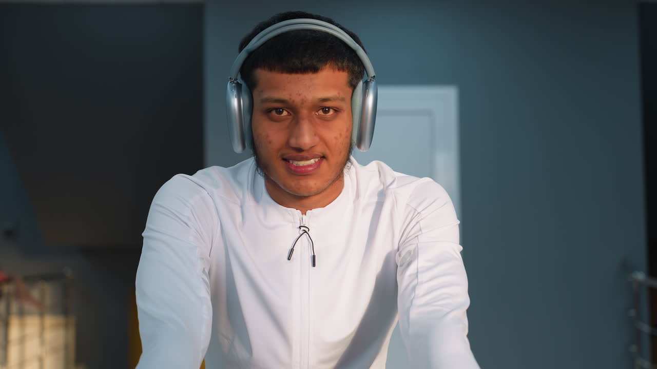 Calm man in white active top and headset cycles on stationary bike with warm smile, blurred hand and gym equipment visible in background under bright ceiling lights
