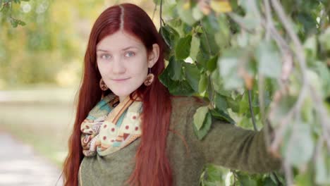 4k cerca de una hermosa joven pelirroja parada en un parque en una soleada tarde de otoño con ramas de árboles colgando a su lado