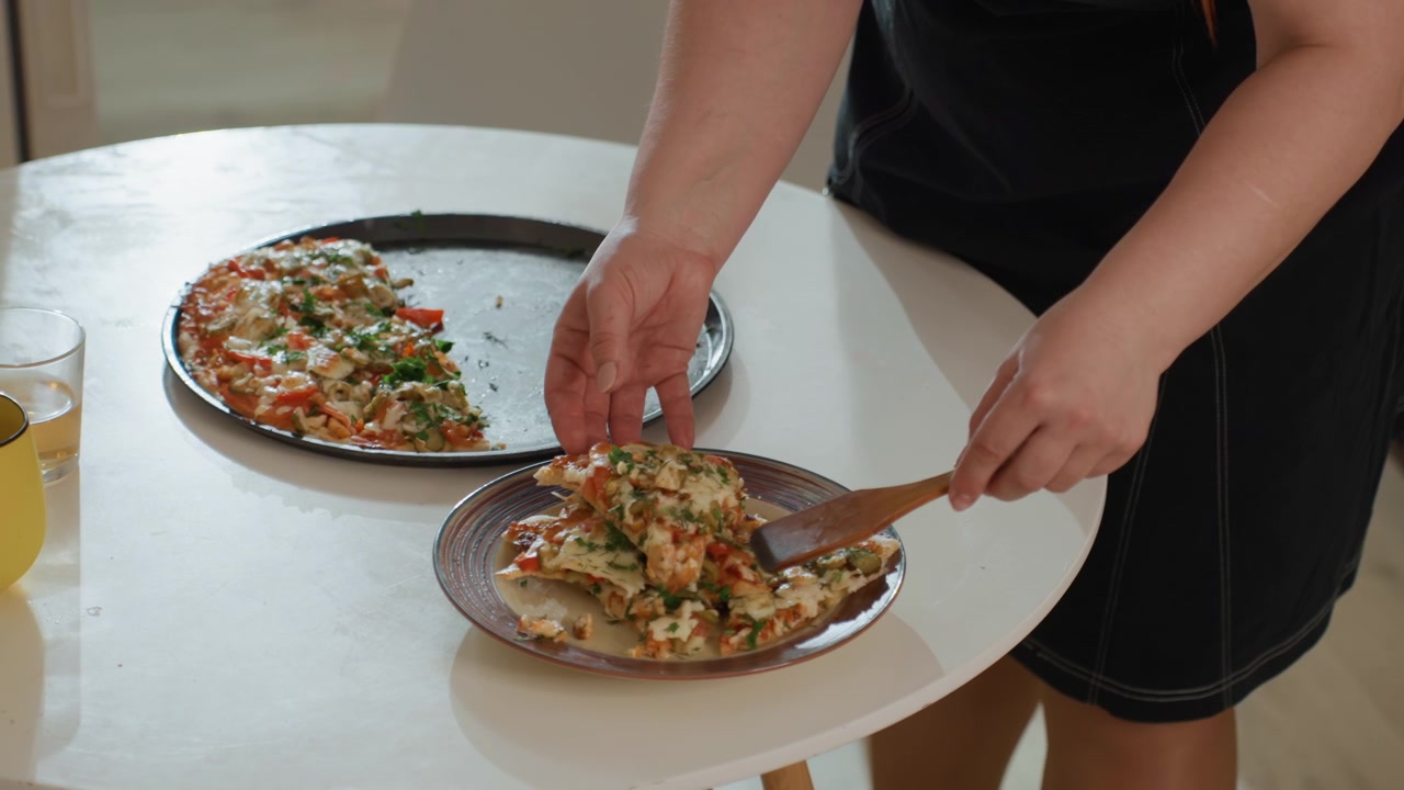 Zbliżenie rąk krojących, podnoszących i układających kawałki pizzy z okrągłej formy na mały ceramiczny talerz na białym stole; widoczne czynności obejmują krojenie nożem, podnoszenie łopatką i stabilizowanie formy podczas