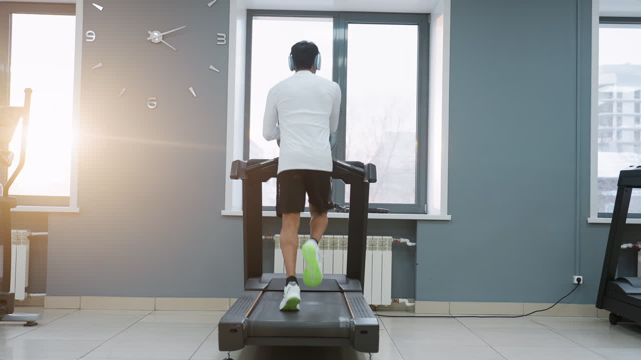 back view of man running on treadmill in bright modern gym interior next to large windows with sunrise glow clock on wall dynamic workout session showcasing calf muscles motion energy