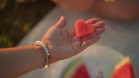 primer plano de la mano sosteniendo una rebanada de sandía en forma de corazón, que simboliza el amor y la frescura, con una delicada pulsera que mejora la elegancia en la cálida luz exterior